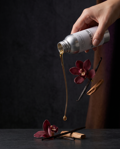 A hand pours a viscous golden amber liquid from a silver bottle labeled &
